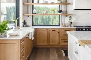 Bright kitchen featuring wooden cabinetry, white farmhouse sink, marble countertops, floating wood shelves, and large windows overlooking forest views.
