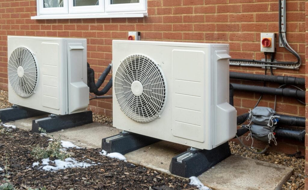 Two white air source heat pump units mounted on a brick exterior wall with black condensate pans and connected piping.