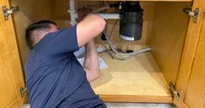 Plumber working under kitchen sink to install or repair PVC drain pipes and garbage disposal system.