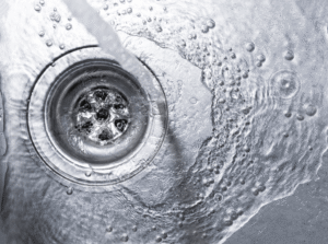 Water flowing down a drain opening with bubbles and water droplets visible on the metallic surface surrounding it.