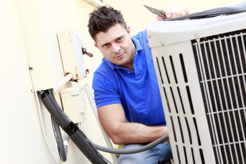 Male technician in blue shirt inspecting an air conditioning unit's outdoor condensing coil and refrigerant lines.